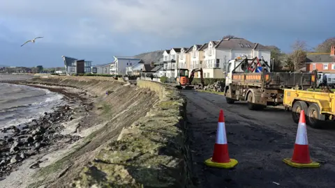 Pacemaker The image shows at Rhanbuoy Park in Carrickergus, a coastal road with the sea on one side and a row of houses on the other. two traffic ocnes are in the foreground, debris can be seen on the road. A large lorry and a sammler digger are on the road.