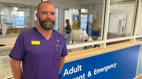 James Grant/BBC A man in a purple NHS uniform stands by a reception area. He has a beard and is smiling at the camera.
