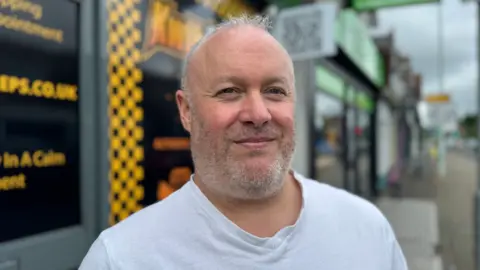 Simon Lawrence standing in front of his shop which has yellow writing and logos. he's got grey hair and wearing a white t shirt