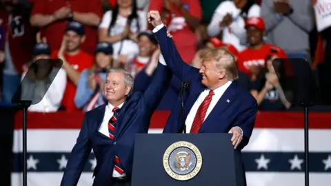 President Donald Trump and Senator Lindsey Graham are wearing navy blue business suits and standing behind a podium with the US presidential seal holding, each holding an arm up toward the other in a victorious gesture