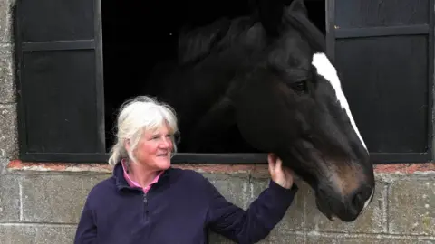 John Fairhall/BBC Ms Lock stands outside a stable. She rests one hand on the underside of Mylo's head as he leans out of the stable door. Ms Lock has grey hair and wears a navy jumper with a pink polo top underneath.