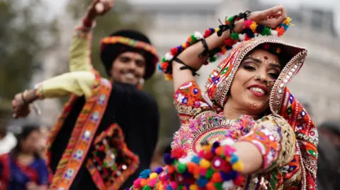 A man dancing on the left in black traditional Indian clothing with a woman on the right with a colourful Indian outfit on also dancing. 