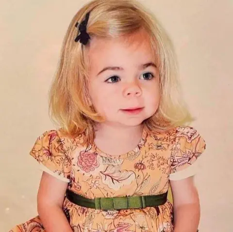 Darren Scott Sophia as a very young child. She has shoulder-length, blonde hair with a black bow clipping it away from her face. She is looking off camera and is wearing an autumnal brown dress with short sleeves and a green bow on a ribbon around it