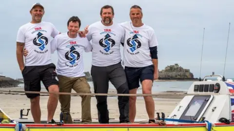 South North Adventures From left to right: rowers Billy Taylor, Prof Kevin Dutton, Gary Hutchings and Patrick Neale smiling for a photo on the shore on their boat on a cloudy day. They are wearing branded T-shirts of their challenge.