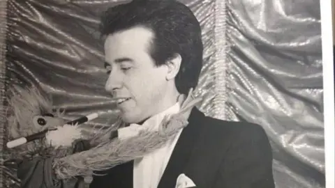 Reg Martin A black and white image of Reg Martin as a younger man performing magic. He is holding a plush doll with a magic wand in its mouth. Reg is looking at the doll.
