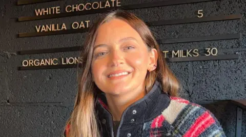 Riot Cycles owner Claire Reynolds standing in front of a black wall with yellow lettering showcasing the prices of her items. She is wearing a woolly cream, red, and black coat. She is smiling looking at the camera.
