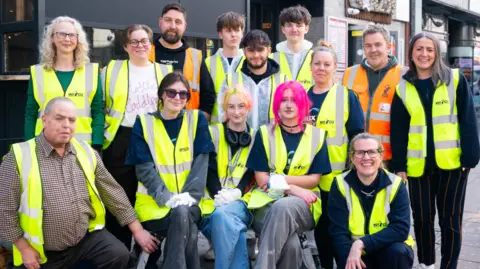 Volunteer It Yourself A group of volunteers outside Voodoo Daddy's in Norwich, wearing fluorescent hi-vis vests. The group is made up of 13 people in the picture. 