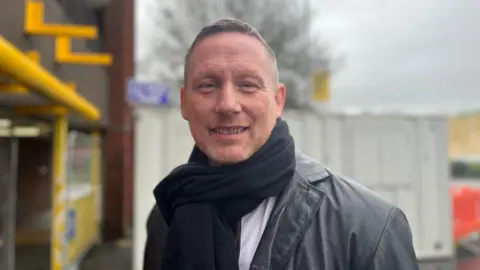 Chris smiles at the camera, standing in front of the closed off entrance to the car park. he wears a black scarf, black jacket and white shirt