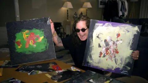A man is wearing a black top and sunglasses while holding a painting in each hand. The painting on the left has a black background and a green middle. The painting on the right is filled with purple and cream brushstrokes.