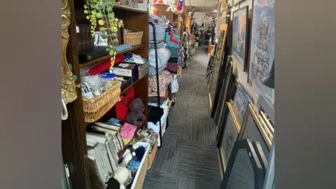 Laura Foster/BBC A narrow corridor with shelving on one wall stacked with fake plants, bedding, teddies, etc and picture frames and mirrors stacked along the opposite wall.