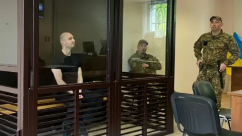 A man in a black t-shirt sits in a witness box guarded by soldiers