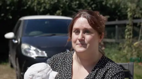 Saffron Alton-Barker wearing a polka dot top. She has her hair tied-up and is holding her baby. There is a black and white car in the background parked on a drive.
