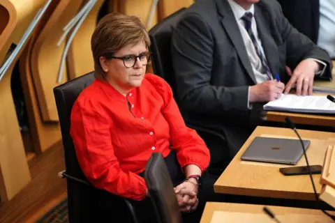 PA Media Nicola Sturgeon sits in the debating chamber of the Scottish Parliament clasping her hands. She wears a red shirt and black glasses and sits next to a person in a dark suit