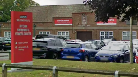 Google A car park in front of a low, brown-brick leisure building with a tiled roof. In the foreground, a large red sign reads “Everyone welcomes you to Hitchin Swimming & Fitness Centre".