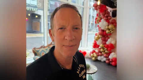 Martin Smith smiling at the camera has grey hair and blue eyes. He is wearing a black polo shirt and is standing in a tattoo shop.