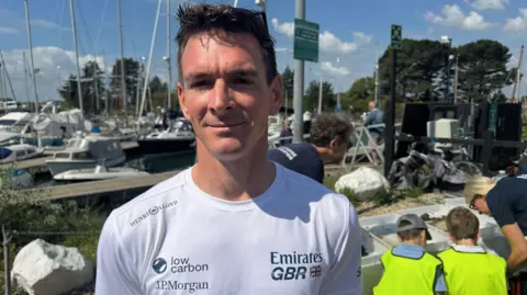 A man with short brown hair looks into the camera. He is wearing a white t-shirt bearing the logos Emirates GBR, low carbon and JP Morgan. Behind him are moored boats and two children with yellow high-vis tabards who are sat with their backs to the camera, leaning over boxes containing items that cannot be made out. A woman with blonde hair and a dark t-shirt leans over next to them, looking at the same things.