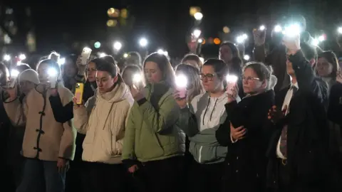 PA A group of women gather with their lights on their phones looking mournful.