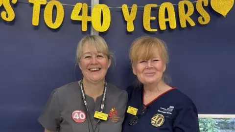 Two nurses with blonde hair stand smiling side by side under a banner which celebrates their 40 years in the job.