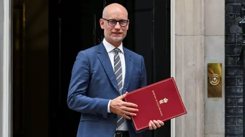 Reuters Stephen Kinnock standing in the doorway of 10 Downing Street with the black door closed behind him. He is bald with a short grey beard and moustache and is wearing glasses and dressed in a blue suit, white shirt and grey striped tie. he person is holding a large red ministerial-style folder or document case.
Gold lettering is visible on the folder, including the words "SECRETARY OF STATE FOR WALES" and a royal insignia. There is the hint of a smile on his face.