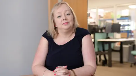 Julia is wearing a dark navy, sleeveless dress. She is sat in her offices, with her hands clasped in front of her, on a desk. She has shoulder length, straight blonde hair. Behind her is an open door to the office, which is slightly blurred.
