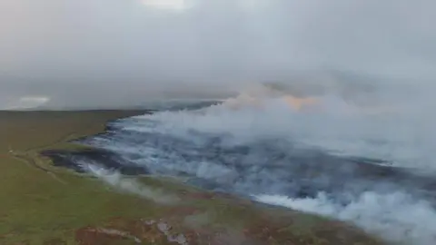South Wales Fire and Rescue Service An image taken from the air shows huge clouds of smoke rising from a wildfire in the near distance. Wind blows it from left to right across the image. 