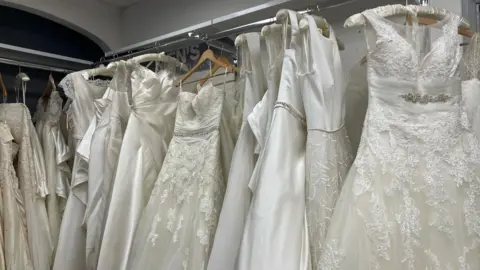 Charlotte Walker Picture of a row of wedding dresses in a store. There are around 14 dresses in colours of white and ivory. They are hung upon a silver clothes rail, some are satin while others are flowered lace layered over tulle. 