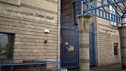 The outside of Wolverhampton Crown Court, with silver lettering on the front of the building and blue metal gates at the entrance.