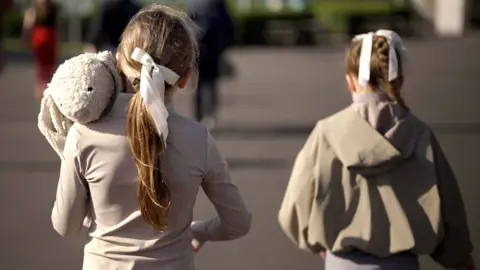 Two young girls walk alongside each other with their backs to the camera. They're wearing grey with their hair tied back in a bow. The one on the left is carrying a large stuffed toy rabbit. 