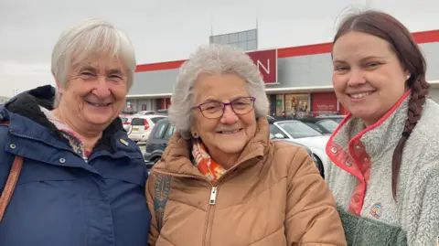 Three happy looking, smiling women; Christine and Anne are older women both with grey hair. Christine wears a blue winter coat, Anne wears purple rimmed glasses, a tan coloured winter puffer style coat with a bright orange and yellow scarf just visible around her neck. Alex too is smiling broadly, and has long brown hair worn in a plait down to one side and a two tone fleece grey at the top and grey/green at the bottom. It also has a rainbow style logo on the left lapel and orange trimmings, and poppers at the neck.
In the background there are cars visible, and part of the signage of the Matalan store visible just above their heads.