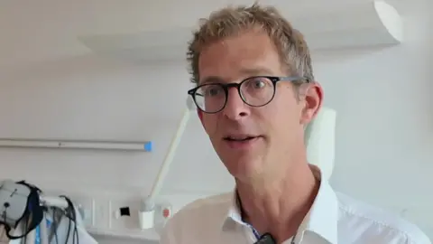 A man with glasses and short greying hair is in a clinical setting talking to someone away from the camera.