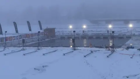 Michelle Leach An outdoor pool, lit with floodlights, in very misty surroundings. There are promotional flags around the pool, and it has been divided into lanes. The pool is surrounded by deep snow on the ground.
