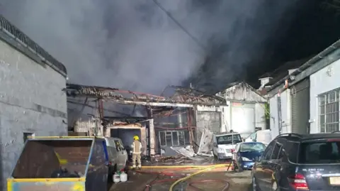 Number of workshop type buildings one has its roof missing after a fire with smoke rising and a fire fighter stands in front of it.