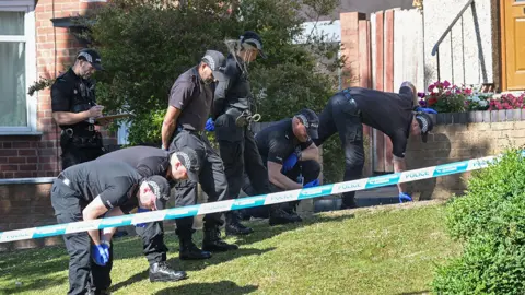 Alamy Seven police officers in black uniforms and baseball-style caps closely inspect a lawn at the scene of a shooting. They seem to be searching it for evidence. One of the officers is behind the others holding a clipboard