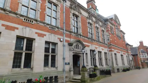 The front entrance to County Buildings in Stafford - a large two-storey building which is half beige and half orange bricks. 