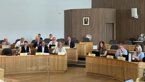 Local Democracy Reporting Service A committee sits inside a council chambers at large wood panelled desks. There are 13 people sat down with some empty chairs at desks behind them.