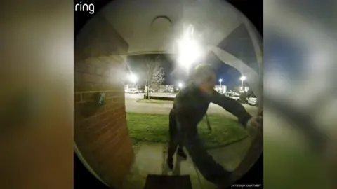 Man wearing dark clothing looking into a house, captured on a Ring doorbell.