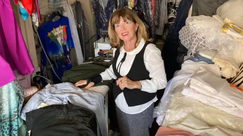 Justin Dealey/BBC A woman stands in a clothes shop surrounded by piles of clothing.