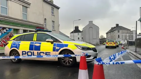 BBC A number of police cars behind a blue and white police cordon on West Hoe Road in Plymouth. 