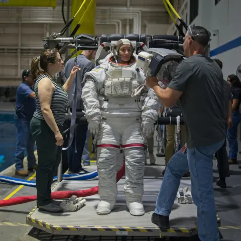 Kevin Church/BBC Astronaut Rosemary Coogan is standing on a platform next to a swimming pool  at the Johnson Space Centre, Houston, Texas. She is wearing a white spacesuit - similar to the ones worn by the Apollo astronauts who landed on the moon. She's wearing a cap on her head that has a microphone attached to it. Two people are helping her. One woman in a striped vest and black trousers and a man in a black T-shirt and blue jeans who is holding the helmet that he is about to put over Rosemary's head. She is looking towards him. There are numerous other people in the background by the side of the pool. 