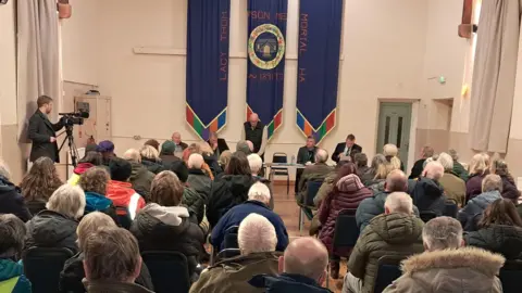 Six rows of people in the cream-coloured hall are facing a panel of five people at the front. Three blue flags hang from the wall behind them and the tall curtains on the side of the room are also cream. There is a man in a black coat operating a camera on the left hand side.