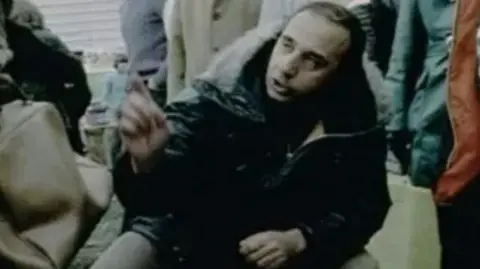 William Goad waving a finger at the camera. He has dark hair and wearing a Parka style anorak in a dated image