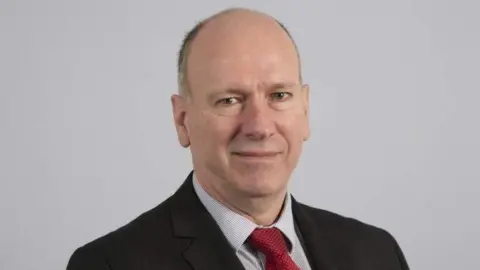 David Rees stood in front of a grey background, wearing a black suit, red tie and shirt.
