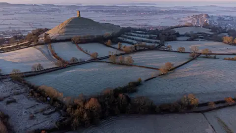 PA Media A wide shot of the Somerset Levels at dawn with Glastonbury Tor in the centre and fields around it covered in frost