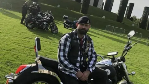 Anthony Frith Anthony Frith sits on a motorbike at the National Memorail Arboretum. He is wearing a beret, a black and white checked shirt and sleeveless jacket which has medals on it. In the backgroud is the Armed Forces Memorial and two motorbikes and several other riders.