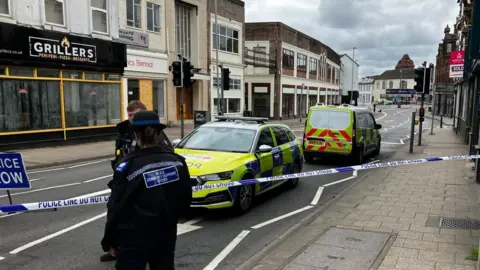 BBC A high street with a police cordon across. Two police vehicles are parked behind the cordon and two officers and standing in front of it.