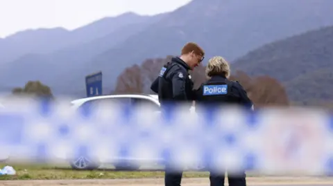 Two police officers stand behind police tape in Porepunkah, green mountains can be seen in the background
