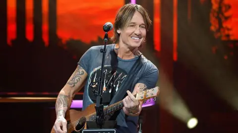 Keith Urban is playing on stage. He has brown hair down past his ears and is wearing a grey/blue T-shirt and playing an acoustic guitar and smiling. Behind him the red/black backdrop of the stage can be seen, in front of him is a microphone on a stand.