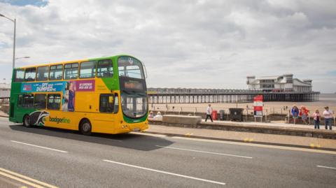 A yellow and green bus passes a beach, with a pier in view in the background