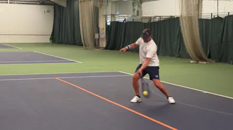 Neil Bamford is swinging his racket to hit the tennis ball during a game in this action shot. He is wearing a white tennis T-shirt, black shorts, white trainers and a black visor.