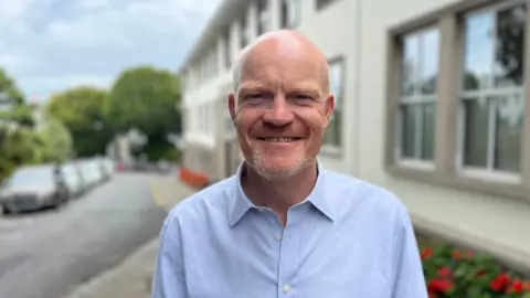 A bald man smiling, wearing a blue shirt. There is a road and buildings in the background.